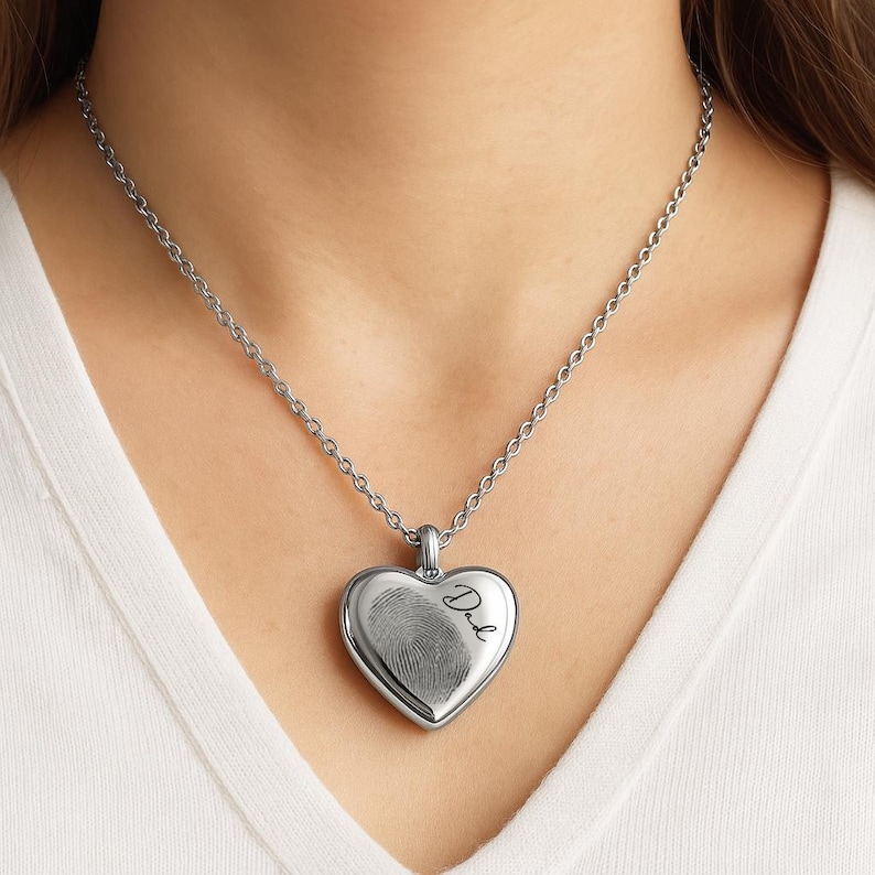 May include: A silver-toned heart-shaped pendant necklace featuring a detailed fingerprint engraving on the front surface. The word Dad is inscribed in a cursive script next to the fingerprint. The pendant hangs from a matching silver-toned chain.