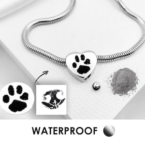 May include: A silver-toned snake chain necklace featuring a heart-shaped pendant engraved with a black paw print. The pendant includes a small screw-in plug for holding ashes, shown next to a pile of grey ash. The text WATERPROOF is printed below.
