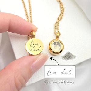 Könnte beinhalten: Goldfarbene Gedenkkette mit rundem Anhänger. Ein Anhänger ist mit "love, dad" in Schreibschrift graviert. Der andere Anhänger hat ein klares Fach. Ein kleiner Aschehaufen befindet sich neben dem Anhänger. Das Bild enthält den Text "Your own handwriting."