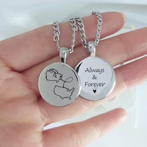 May include: Two silver-toned circular pendant necklaces on metal chains. One pendant features a black engraved map of Canada and the United States connected by a dashed line. The other pendant displays the black text Always & Forever with a small heart.
