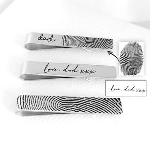 May include: Three silver-toned metal tie clips. The top clip features the word dad and a fingerprint. The middle clip displays the text love, dad xxx. The bottom clip shows a large fingerprint. Two inset boxes show the fingerprint and text details.