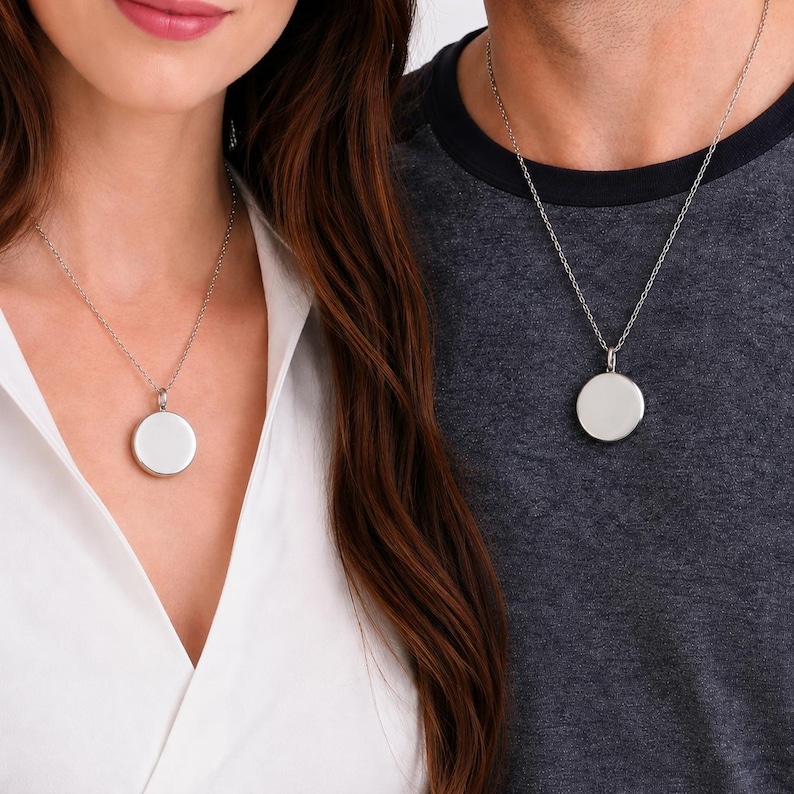 May include: Two silver-toned necklaces featuring a simple, circular pendant suspended from a delicate chain. The pendants are smooth, polished, and round, hanging at the center of the chest. The necklaces are worn over a white shirt and a dark gray shirt.