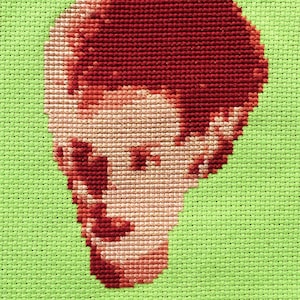 May include: Cross-stitch portrait of a person with red hair and a light pink face. The person is looking down and to the left. The background is a light green.