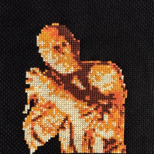 May include: Cross-stitch embroidery of a person in orange and white, against a black background.