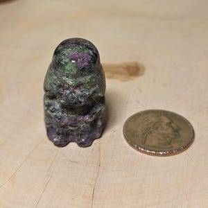 May include: A carved stone figurine with a dark green and purple color scheme. The figurine is next to a US quarter coin. The stone has a rough texture and appears to be a small statue or decorative item.