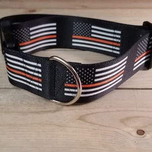 May include: A black dog collar with a thin red stripe and an American flag pattern. The collar has a silver D-ring.