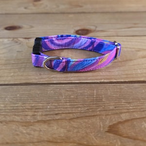 May include: A vibrant pet collar with a colorful, swirling pattern in shades of purple, blue, and pink. The collar features a black plastic buckle and a silver metal D-ring. The collar is designed for small pets.