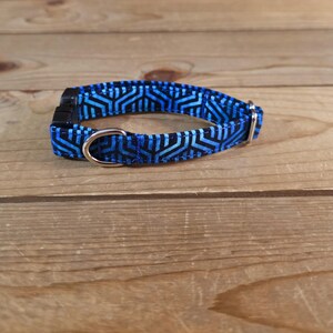 May include: A blue and black patterned dog collar with a silver metal D-ring and a black plastic buckle. The collar is on a wooden surface.