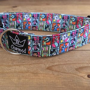 May include: A colorful dog collar with a cartoon character pattern. The collar features a black buckle and a silver metal ring. The brand name "Charmed" is printed on a black label. The collar is on a wooden surface.