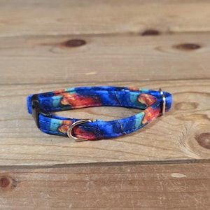 May include: A blue pet collar with a colorful floral pattern in shades of orange, red, and teal. The collar has a black plastic buckle and a gold-colored metal loop. The collar is lying on a wooden surface.