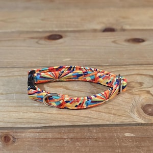 May include: A colorful dog collar with a geometric pattern in shades of orange, yellow, blue, and red. The collar has a black buckle and a silver metal loop for attaching a leash. The collar is made of fabric.