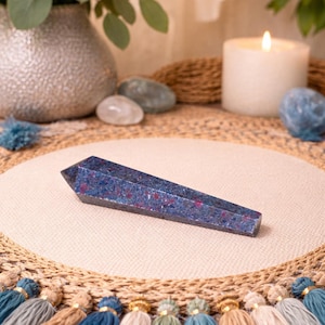 May include: A blue and red crystal obelisk, a decorative item, rests on a beige surface. The obelisk is surrounded by a candle, stones, and a vase with greenery. Tassels in various colors are at the bottom.