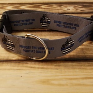 May include: Grey dog collar with a thin blue line American flag paw print design and the text "Support the paws that protect our laws".