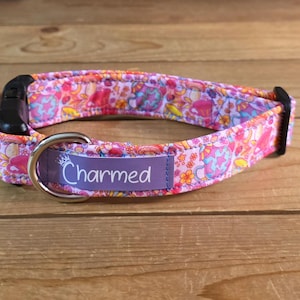 Pode incluir: Uma coleira rosa para cães com um padrão floral e um anel de metal prateado. A coleira tem uma etiqueta roxa com a palavra "Charmed" em escrita branca. A coleira é feita de tecido e tem uma fivela de plástico preta.