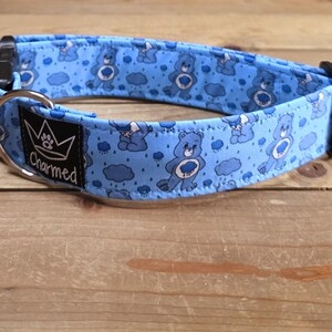 May include: A light blue pet collar featuring a repeating pattern of blue cartoon bears and rain clouds. The collar includes a black plastic side-release buckle, a silver metal D-ring, and a black fabric tag with a crown logo and the text Charmed.