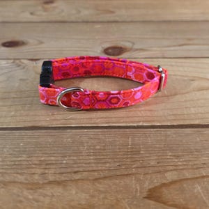 May include: A brightly coloured dog collar with a geometric pattern in shades of red and pink. The collar has a black plastic buckle and a silver metal D-ring. The collar is made of fabric and is designed for pets.
