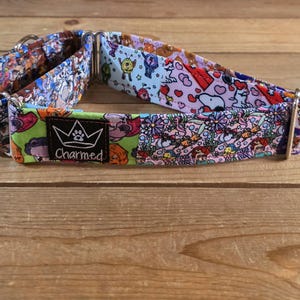 May include: A colorful dog collar with various cartoon character patterns. The collar features a black label with a crown logo and the word "Charmed". The collar is made of fabric and has metal hardware.