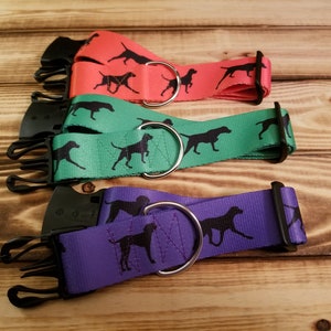 Può includere: Tre collari per cani in diversi colori: rosso, verde e viola. Ogni collare ha una silhouette nera di un cane stampata su di esso. I collari hanno fibbie in metallo nero e anelli a D in metallo argentato.