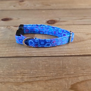 May include: A blue pet collar with a geometric pattern in shades of blue and purple. The collar has a black plastic buckle and a silver metal D-ring. The collar is approximately 1.3 cm wide.