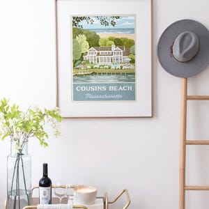 May include: Framed art print featuring a coastal scene with a large house, trees, and beach. The print is labeled "Cousins Beach Massachusetts" in a light blue banner. A gray hat hangs on a wooden ladder, and a bar cart with wine and glasses is in the foreground.