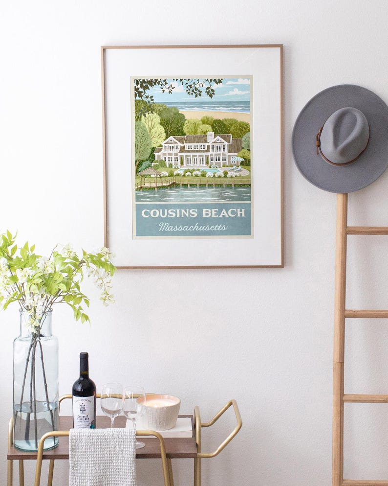 May include: Framed art print featuring a coastal scene with a large house, trees, and beach. The print is labeled "Cousins Beach Massachusetts" in a light blue banner. A gray hat hangs on a wooden ladder, and a bar cart with wine and glasses is in the foreground.