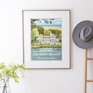 May include: Framed art print featuring a coastal scene with a large white house, trees, and a beach. The print includes the text "COUSINS BEACH Massachusetts" in a blue banner. A gray hat hangs on a wooden rack to the right.