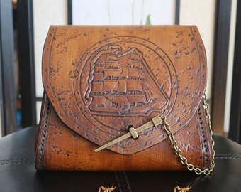 May include: A brown leather purse with a tooled design of a ship on the front. The purse has a brass clasp and a chain strap.