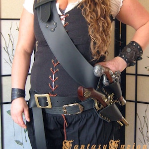 Puede incluir: Una mujer con un sombrero de pirata marrón con una pluma roja, un chaleco de cuero negro con un frente de encaje, un cinturón de cuero negro con una hebilla de latón y una funda de cuero negro con una pistola plateada. También lleva una muñequera de cuero negro con un diseño plateado.