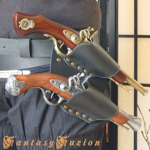 May include: Two antique-style pistols with ornate metalwork and wooden grips are holstered in a black leather double holster with metal studs. The holsters are worn on a black belt.