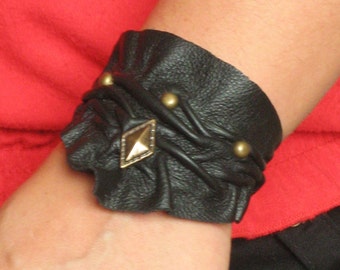 Könnte beinhalten: Schwarzes Lederarmband mit geflochtenem Design und goldfarbenen Akzenten. Das Armband hat ein diamantförmiges Stück in der Mitte.