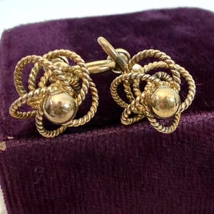 以下が含まれることがあります： 金色のイヤリングのペア。各イヤリングは、ロープのような織り模様で囲まれた中央の球体を特徴としています。イヤリングは、濃い紫色のベルベットの表面に表示されています。