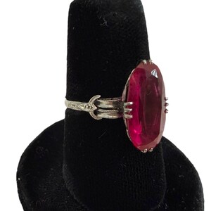 May include: A silver ring with an oval, deep red gemstone. The ring is set on a black display stand. The gemstone is the focal point, showcasing its rich color and shape. The ring's band has an ornate design.