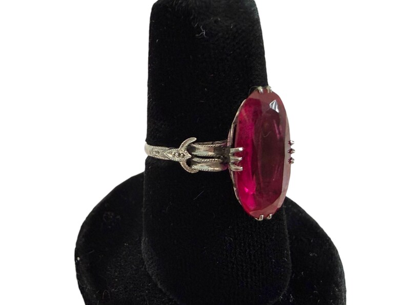 May include: A silver ring with an oval, deep red gemstone. The ring is set on a black display stand. The gemstone is the focal point, showcasing its rich color and shape. The ring's band has an ornate design.