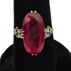May include: A silver ring featuring a large, oval-cut, deep red gemstone. The gemstone is the focal point, set in a decorative silver band with ornate side details. The ring is displayed on a black surface.