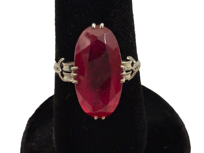 May include: A silver ring featuring a large, oval-cut, deep red gemstone. The gemstone is the focal point, set in a decorative silver band with ornate side details. The ring is displayed on a black surface.