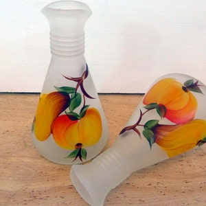 May include: Two frosted glass vases with a hand-painted fruit design. The vases feature a pear-shaped body and a flared top. The fruit is painted in shades of yellow, orange, and red, with green leaves and brown stems.