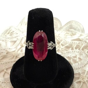 May include: A silver ring featuring a large, oval-cut, deep red gemstone. The gemstone is the focal point, set in a silver band with decorative filigree detailing on each side. The ring is displayed on a black velvet ring holder.