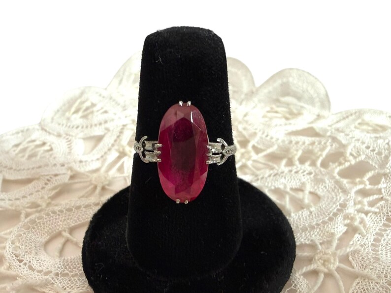 May include: A silver ring featuring a large, oval-cut, deep red gemstone. The gemstone is the focal point, set in a silver band with decorative filigree detailing on each side. The ring is displayed on a black velvet ring holder.