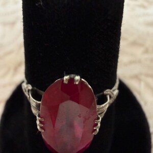 May include: A silver ring featuring a large, oval-cut, deep red gemstone. The gemstone is the focal point, set in a detailed silver band with intricate designs. The ring is displayed on a black velvet surface.