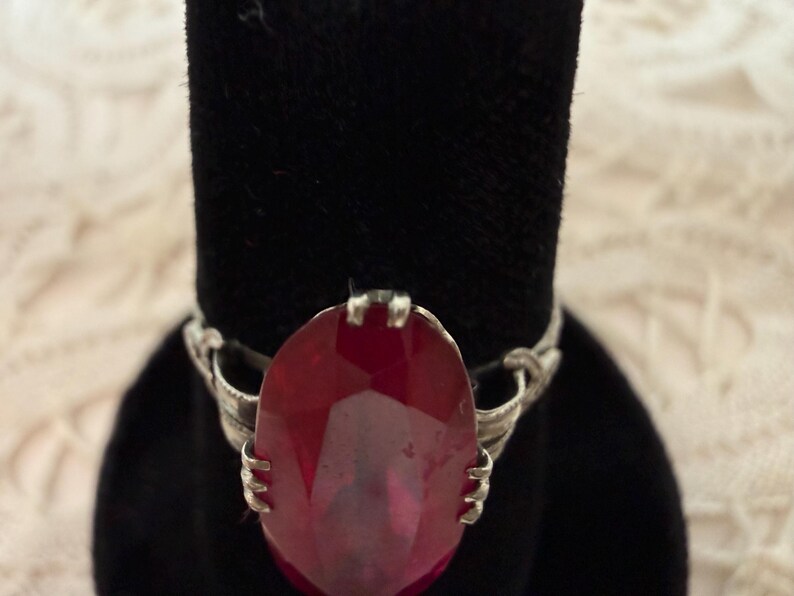 May include: A silver ring featuring a large, oval-cut, deep red gemstone. The gemstone is the focal point, set in a detailed silver band with intricate designs. The ring is displayed on a black velvet surface.