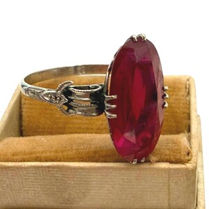 May include: A vintage silver ring featuring a large, faceted, oval-cut ruby gemstone. The ruby is a deep red color and is set in a detailed silver band with ornate filigree designs. The ring is displayed in a small, tan jewelry box.