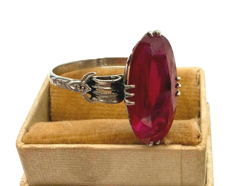 May include: A vintage silver ring featuring a large, faceted, oval-cut ruby gemstone. The ruby is a deep red color and is set in a detailed silver band with ornate filigree designs. The ring is displayed in a small, tan jewelry box.