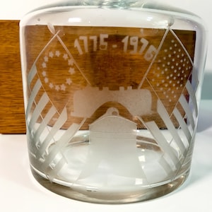 May include: Clear glass decanter with etched design featuring American flags, the Liberty Bell, and the text "1775-1975". The decanter has a wide base and a slightly tapered body. A wooden block is visible on the left side.