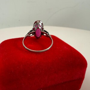 May include: A silver ring with an oval, bright pink gemstone. The ring is set on a red velvet surface. The ring's design includes decorative filigree on the sides of the gemstone setting.