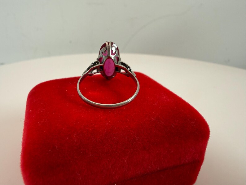 May include: A silver ring with an oval, bright pink gemstone. The ring is set on a red velvet surface. The ring's design includes decorative filigree on the sides of the gemstone setting.