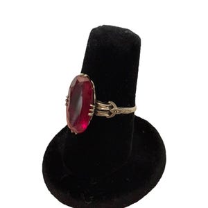 May include: A gold-toned ring featuring a large, oval-cut, deep red gemstone. The ring's band has an ornate design with delicate detailing. The gemstone is the focal point, set in a classic, elegant style. The ring is displayed on a black velvet ring holder.