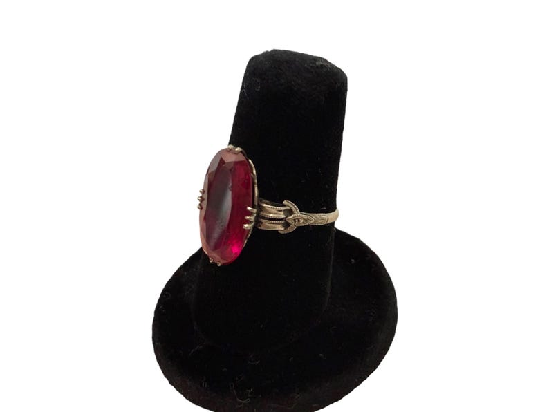 May include: A gold-toned ring featuring a large, oval-cut, deep red gemstone. The ring's band has an ornate design with delicate detailing. The gemstone is the focal point, set in a classic, elegant style. The ring is displayed on a black velvet ring holder.