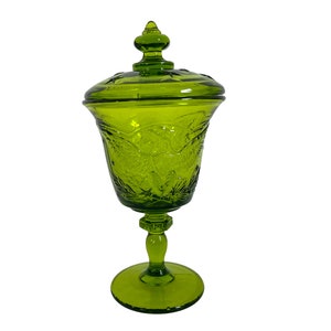 May include: A green glass candy dish with a lid. The dish has a pedestal base and is decorated with floral patterns. The lid has a finial on top. The glass is a vibrant shade of green.