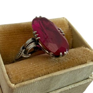 May include: A silver ring with a large, faceted, oval-shaped, red gemstone. The ring is set in a decorative silver band and rests in a vintage, beige velvet-lined ring box. The ring box is a light beige color.