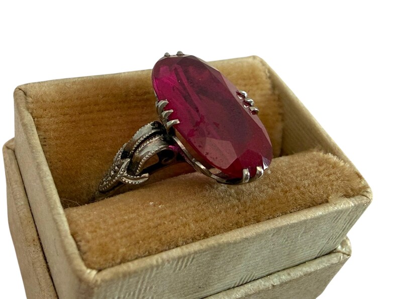 May include: A silver ring with a large, faceted, oval-shaped, red gemstone. The ring is set in a decorative silver band and rests in a vintage, beige velvet-lined ring box. The ring box is a light beige color.